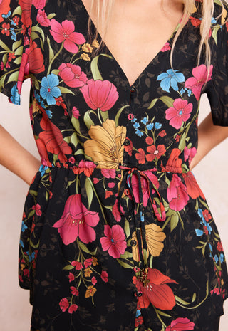 a-close-up-of-a-black-short-sleeve-v-neck-dress-with-a-vibrant-floral-print-featuring-pink-red-blue-and-yellow-flowers-with-green-leaves-the-dress-has-a-drawstring-waist-and-button-details-down-the-front